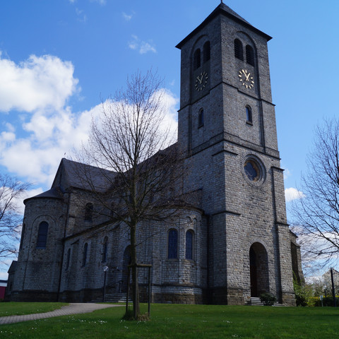 Kirche "Sankt Johannes der Täufer" in Uckerath Eine historische Kirche mit eindrucksvollem Glockenturm aus grauem Stein vor blauem Himmel.