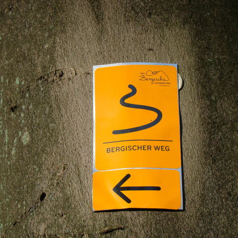 Markierungszeichen Bergischer Weg Wanderweg-Markierung am Baum für den Bergischen Weg, gelbes Schild mit schwarzem Pfeil und Serpentine.