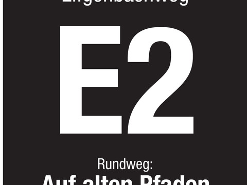 Markierungszeichen E2 Schwarz-weißes Schild mit der Aufschrift "Eifgenbachweg E2 Rundweg: Auf alten Pfaden" in weißer Schrift.