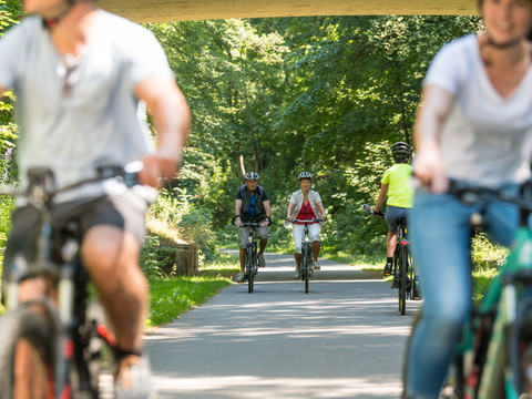 Balkantrasse Radfahrer auf einem Waldweg unter einer Brücke, im Vordergrund unscharf, sonniger Tag.