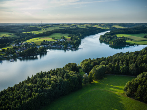 Bevertalsperre Luftbild eines weitläufigen Sees, umgeben von Wäldern, Wiesen und kleinen Siedlungen unter blauem Himmel.