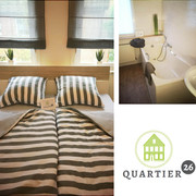 fewo-quartier26 Doppelbett mit gestreifter Bettwäsche in hellem Raum, daneben Badezimmer mit moderner Badewanne.