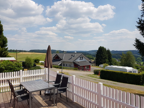 Büschem's Box Terrasse mit Tisch und Stühlen, mit Blick auf Hügel, Felder und Häuser unter blauem Himmel.