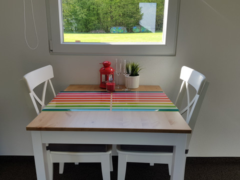 Büschem's Box Ein einladender Holztisch mit buntem Tischläufer, weißen Stühlen, Kerze und Pflanze vor Fenster.