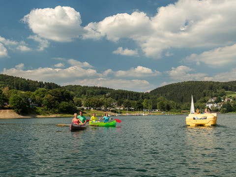 Freizeitcamp Aggertalsperre <p>Menschen paddeln in bunten Kanus auf einem See, umgeben von grünen Hügeln und blauem Himmel.</p>