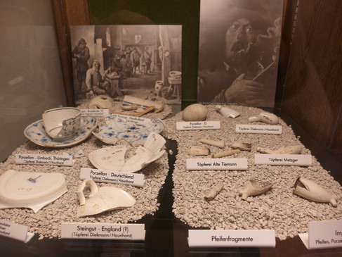 Porzellanscherben und Pfeifenfragmente in einer Vitrine auf Kieselsteinen ausgestellt.