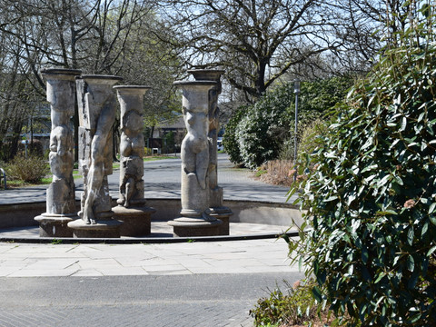 Kurpark in Nümbrecht  Skulpturengruppe aus Steinsäulen mit Reliefs auf einem Platz, umgeben von Bäumen und Sträuchern.