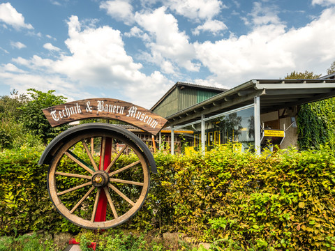 Technik- und Bauernmuseum Das Technik- und Bauernmuseum in Much mit großem alten Mühlrad vor bewachsener Fassade und blauem Himmel.