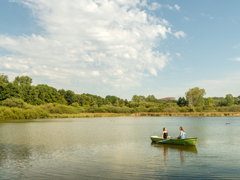 Herrenteich Zwei Personen sitzen in einem Ruderboot auf einem ruhigen See, umgeben von grüner Natur.
