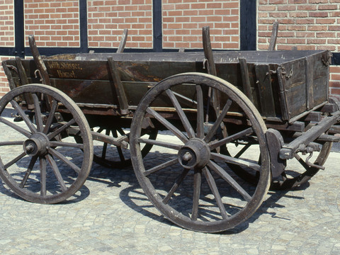 Fuhrmannswagen Ein alter, hölzerner Pferdewagen mit vier großen Rädern steht auf einem Kopfsteinpflaster vor einer Backsteinmauer.