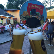 Die Alteburg Zwei Kölsch Gläser stoßen an und im Hintergrund sitzen Menschen im Innenhof.Two Kölsch glasses clink and in the background, people are sitting in the courtyard.