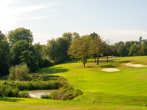 Golfplatz Burg Overbach Eine sanfte Golfplatzlandschaft mit Teich, Bäumen und Sandbunkern unter blauem Himmel.