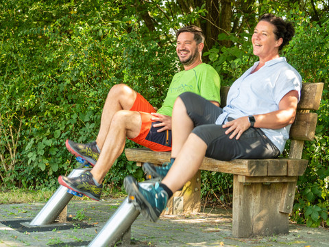 Kneipp-Vitalpark Zwei Personen lachen und trainieren auf einer Bank im Freien, umgeben von grünen Büschen.