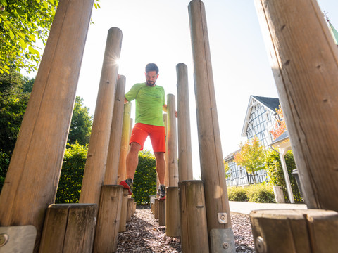 Kneipp-Vitalpark <p>Ein Mann balanciert auf Holzbalken in einem sonnigen Park mit Fachwerkhäusern im Hintergrund.</p>