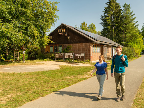 Aggerbogen Zwei Personen spazieren auf einem sonnigen Weg vor einem Backsteingebäude in einer grünen Landschaft.