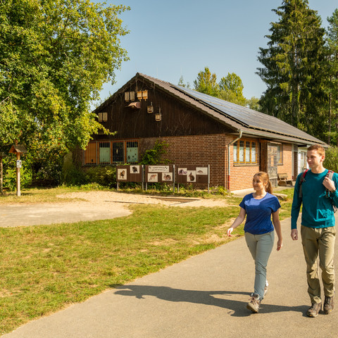 Aggerbogen Zwei Personen spazieren auf einem sonnigen Weg vor einem Backsteingebäude in einer grünen Landschaft.