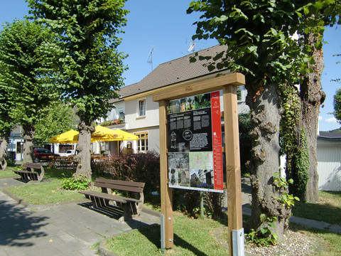 Infotafel am Steinhauerpfad Ein Infobrett neben Parkbänken und Bäumen, im Hintergrund ein Gebäude mit gelben Sonnenschirmen.