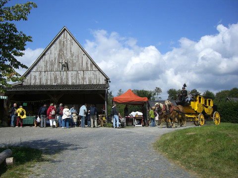Bauernmarkt im Freilichtmuseum  <p>Traditionelle Postkutsche vor historischem Fachwerkgebäude mit Besuchern auf steinigem Platz.</p>