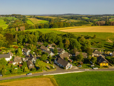 Lohmar-Muchensiefen  Luftaufnahme eines idyllischen Dorfes, umgeben von grünen Feldern und Wäldern, unter blauem Himmel.