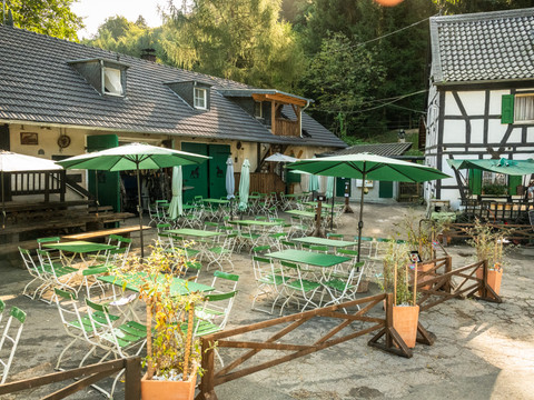 Gammersbacher Mühle <p>Grüne Tische und Sonnenschirme auf einem ruhigen Hof mit Fachwerkhaus und Bäumen im Hintergrund.</p>
