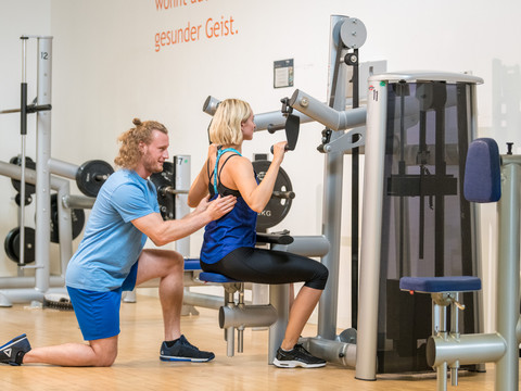 Trans World Hotel Kranichhöhe Eine Frau trainiert im Fitnessstudio an einem Kraftgerät, während ein Trainer sie unterstützt.