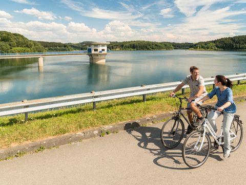 Radfahren an der Dhünntalsperre Radfahrerpaar am sonnigen Stausee, von Wäldern umgeben, unter blauem Himmel mit weißen Wolken.