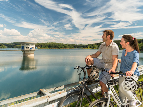 Radfahren an der Dhünntalsperre Ein Paar auf Fahrrädern genießt den Blick auf eine ruhige Stausee-Landschaft an einem sonnigen Tag.