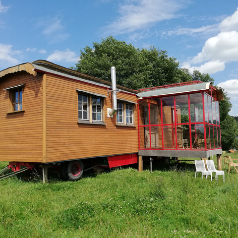 Außenansicht 2 Ein hölzerner Zirkuswagen mit verglastem Anbau steht auf einer grünen Wiese unter blauem Himmel.