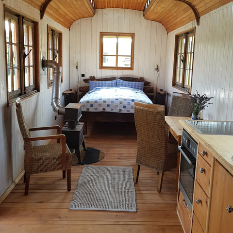 Innenansicht Innenraum eines gemütlich eingerichteten, hölzernen Wagens mit Bett, Sitzbereich und Küchenzeile.