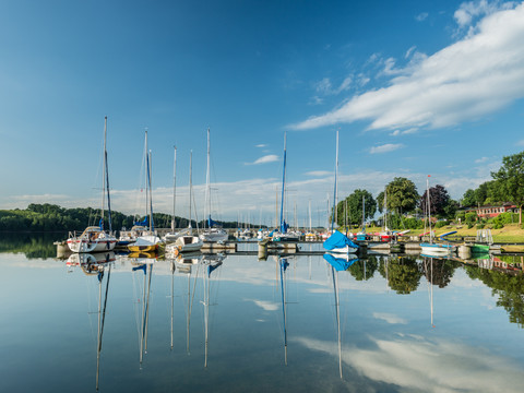 Bevertalsperre Segelboote im ruhigen Wasser, spiegeln sich unter blauem Himmel und weißen Wolken im Hafen wider.