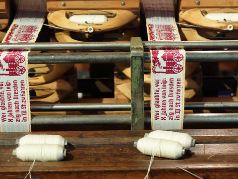 Webstuhl mit traditionellem Muster in Rot und Weiß, Fäden im Vordergrund. Text "Wir glauben...".Loom with traditional pattern in red and white, threads in the foreground. Text "We believe...".Tkalcovský stav s tradičním červenobílým vzorem, nitě v popředí. Text "Věříme...".Krosno z tradycyjnym wzorem w kolorze czerwonym i białym, nici na pierwszym planie. Tekst "Wierzymy...".Weefgetouw met traditioneel patroon in rood en wit, draden op de voorgrond. Tekst "Wij geloven...".Telaio con motivo tradizionale in rosso e bianco, fili in primo piano. Testo "Noi crediamo...".