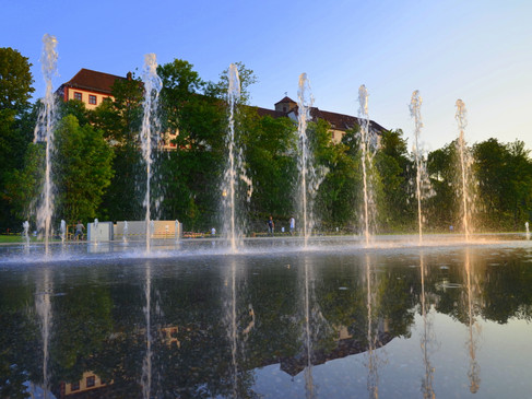 Der Kneipp-Erlebnispark in Bad Iburg am Abend Springbrunnen mit Wasserspielen und Spiegelung, Bäume und Gebäude im Hintergrund bei Sonnenuntergang.Fountain with water features and reflection, trees and buildings in the background at sunset.Springvand med vandfunktioner og refleksion, træer og bygninger i baggrunden ved solnedgang.Fontein met waterpartijen en weerspiegeling, bomen en gebouwen op de achtergrond bij zonsondergang.