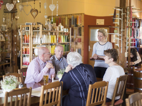 Krietzschwitzer Kerzenträume Eine Gruppe älterer Menschen sitzt an einem Holztisch in einem gemütlichen Laden mit bunten Kerzen im Hintergrund, während eine Frau sie bedient.