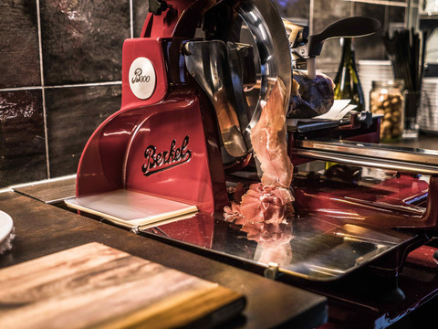 Rote Berkel Aufschnittmaschine schneidet dünne Scheiben Schinken; im Hintergrund Fliesenwand und Küchenutensilien.