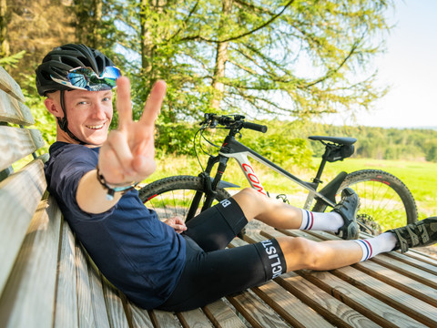 Mountainbike Ein entspannter Radfahrer macht ein Peace-Zeichen auf einer Bank mit seinem Fahrrad im Hintergrund.