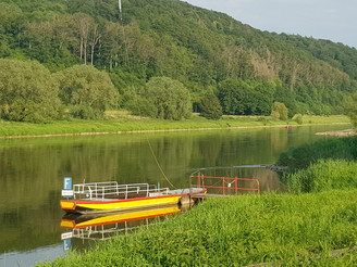 5a_hx_zbih_gierseilfaehrewehrden_stephanberg.jpg Eine gelbe Fähre ankert an einem grünen Ufer vor einem bewaldeten Hang, im ruhigen Flusswasser.