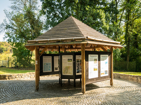 Wahnbachtalsperre Runder hölzerner Pavillon mit Infotafeln in einem grünen Park, umgeben von Bäumen und Kopfsteinpflaster.