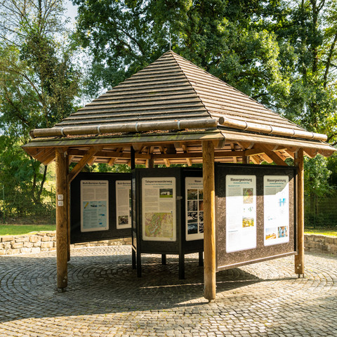 Wahnbachtalsperre Runder hölzerner Pavillon mit Infotafeln in einem grünen Park, umgeben von Bäumen und Kopfsteinpflaster.