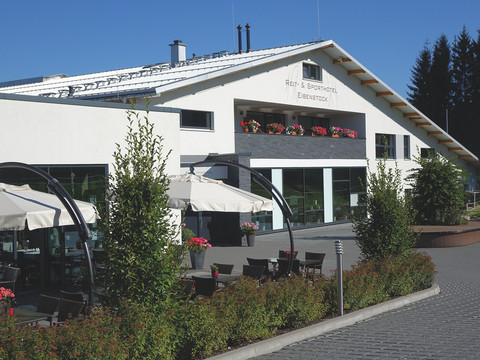 Hotelansicht Helles Hotel mit Sonnenschirmen, Blumen auf Balkonen und umgeben von Bäumen und Sitzgelegenheiten.