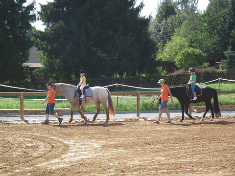 Reitplatz Zwei Kinder reiten auf Pferden, begleitet von Erwachsenen auf einem sandigen Reitplatz.