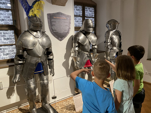 I bambini osservano tre armature di cavalieri medievali in un museo con un pannello informativo.