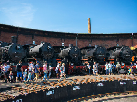 A group of people inspect historic steam locomotives in front of a roundhouse.