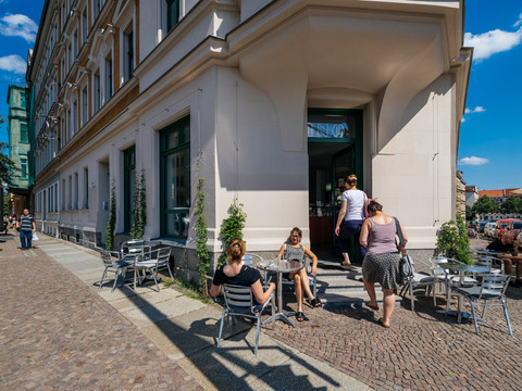 Blick auf den Café-Eingang an der Spitze des Eckhauses sowie den Freisitz vor dem Café mit seinen Besuchern; bei Sonnenschein, freisitz, cafe, gastronomie