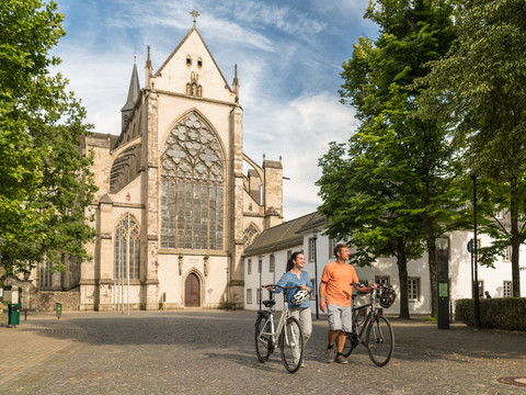 Rad fahren in Altenberg  <p>Ein Paar schiebt Fahrräder vor dem Altenberger Dom bei sonnigem Wetter, umgeben von Bäumen.</p>