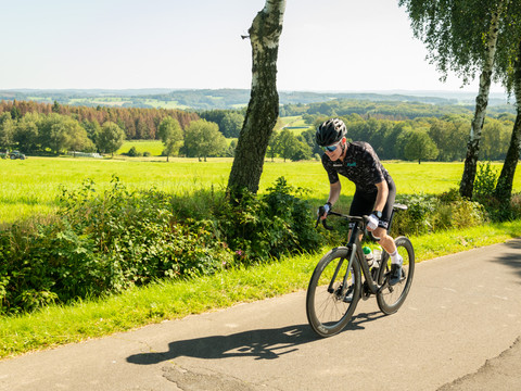 Rennrad <p>Ein Radfahrer in sportlicher Montur fährt auf einer Landstraße durch eine grüne, hügelige Landschaft.</p>