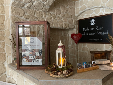 Eingangsbereich Bergwirtschaft Kuhstall Ein Schaukasten mit einem Poster "Wintergeschichten" steht auf einem Steintresen, daneben eine weiße Laterne mit Kerze und ein Herzdekor an der Wand.
