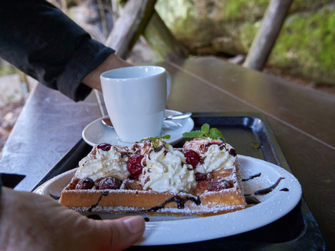 Waffel mit Sahne, Kirschen und Schokoladensauce auf einem Teller, daneben eine weiße Tasse auf einem Tablett, im Hintergrund ein rustikaler Holztisch.