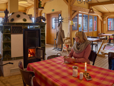 Gastraum Brand-Baude Eine Frau sitzt an einem Tisch mit rot-kariertem Tischtuch in einem gemütlichen Raum, neben einem brennenden Ofen, und hält eine Tasse.A woman sits at a table with a red-checked tablecloth in a cozy room, next to a burning stove, holding a cup.Žena sedí u stolu s červeným kostkovaným ubrusem v útulné místnosti u hořících kamen a drží v ruce šálek.Kobieta siedzi przy stole z czerwonym kraciastym obrusem w przytulnym pokoju, obok płonącego pieca, trzymając filiżankę.Een vrouw zit aan een tafel met een rood geruit tafelkleed in een gezellige kamer, naast een brandende kachel, en houdt een kopje vast.Una donna siede a un tavolo con una tovaglia a scacchi rossi in una stanza accogliente, accanto a una stufa accesa, con in mano una tazza.