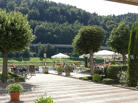 Gepflegte Terrasse mit Tischen und weißen Sonnenschirmen, umgeben von grünen Bäumen und Pflanzen, im Hintergrund ein bewaldeter Hügel.Well-kept terrace with tables and white parasols, surrounded by green trees and plants, with a wooded hill in the background.Udržovaná terasa se stoly a bílými slunečníky, obklopená zelenými stromy a rostlinami, se zalesněným kopcem v pozadí.Zadbany taras ze stolikami i białymi parasolami, otoczony zielonymi drzewami i roślinami, z zalesionym wzgórzem w tle.Verzorgd terras met tafels en witte parasols, omringd door groene bomen en planten, met een beboste heuvel op de achtergrond.Terrazza ben curata con tavoli e ombrelloni bianchi, circondata da alberi e piante verdi, con una collina boscosa sullo sfondo.