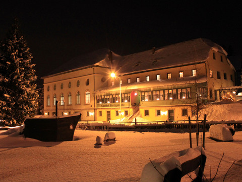 Ein verschneiter Gasthof bei Nacht, beleuchtet von warmem Licht, mit einem großen, schneebedeckten Tannenbaum im Vordergrund.